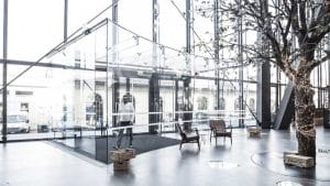 Modern ofis lobisi giriş kapısı, cam duvarlar, ağaç detayı ve rahat oturma alanlarıyla aydınlık bir alan sunuyor.